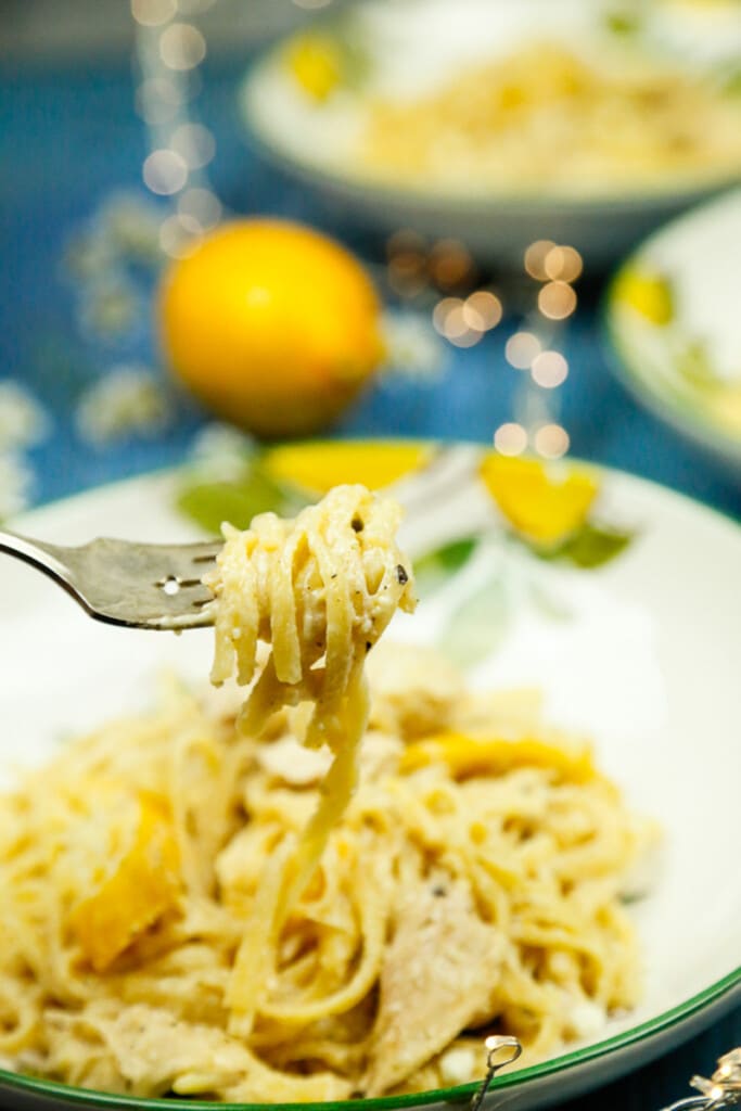 a closeup shot of lemon sauce for chicken and pasta on a fork with a bowl of lemon pasta behind it 