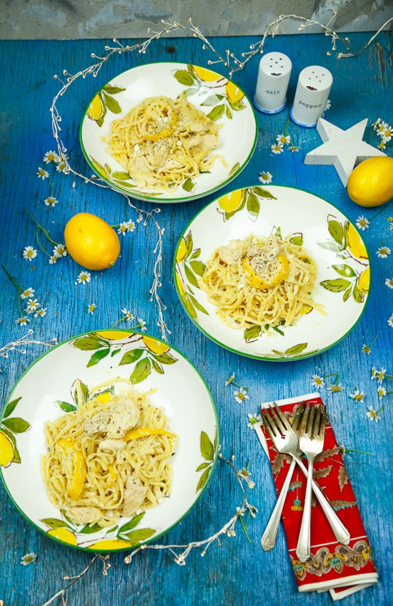 bowls of lemon chicken pasta with lemons and two forks on a blue backdrop 