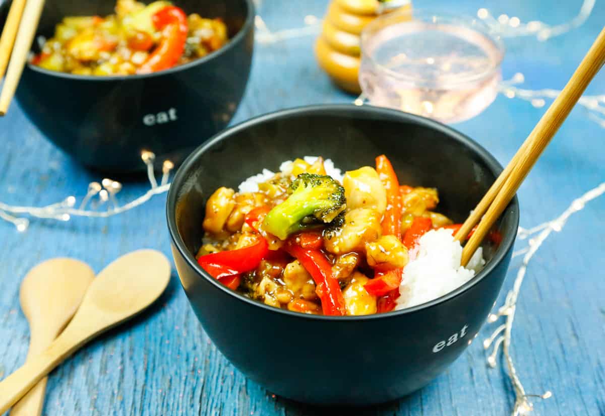 a bowl filled with chicken, rice, veggies, chopsticks with two wooden spoons on a blue wooden backdrop 