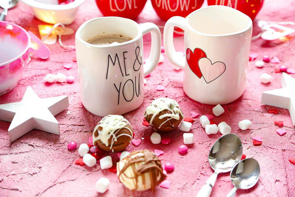 two Rae Dunn mugs with two hearts on them on a pink backdrop with spoons and hot chocolate bombs with white and milk chocolate drizzle