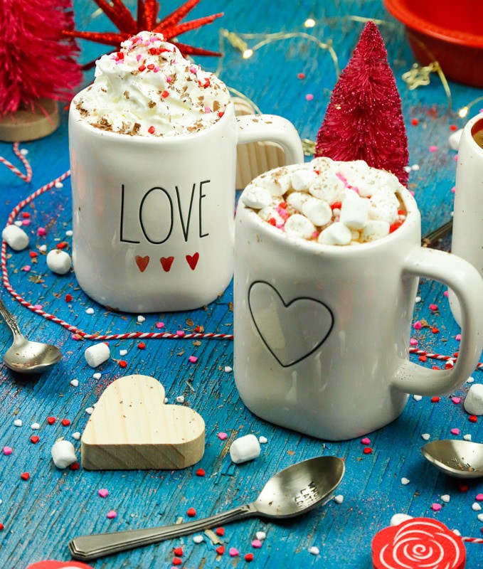 two white Rae Dunn mugs with a black heart on it and the word love with three watercolor hearts filled with cacao hot chocolate and homemade whipped cream and marshmallows 