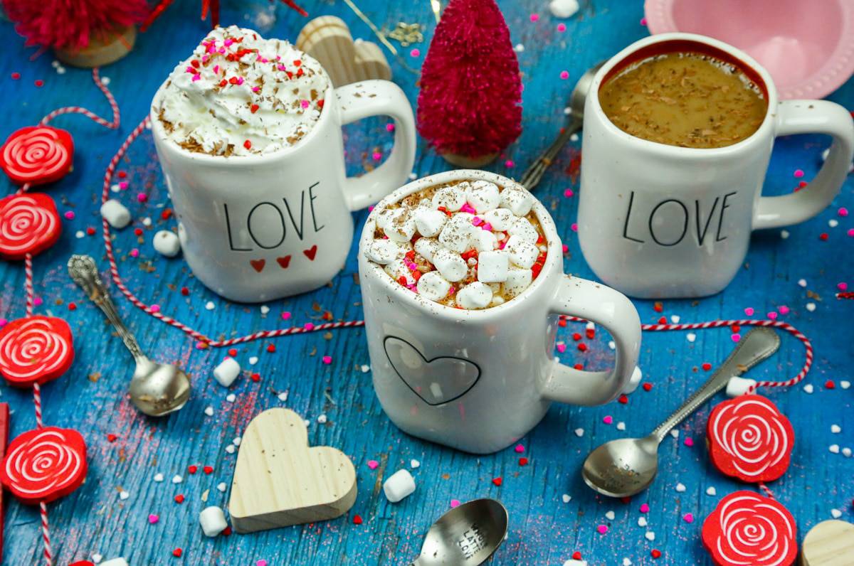Three ceramic mugs of hot chocolate with heavy cream on a blue backdrop with a wooden heart