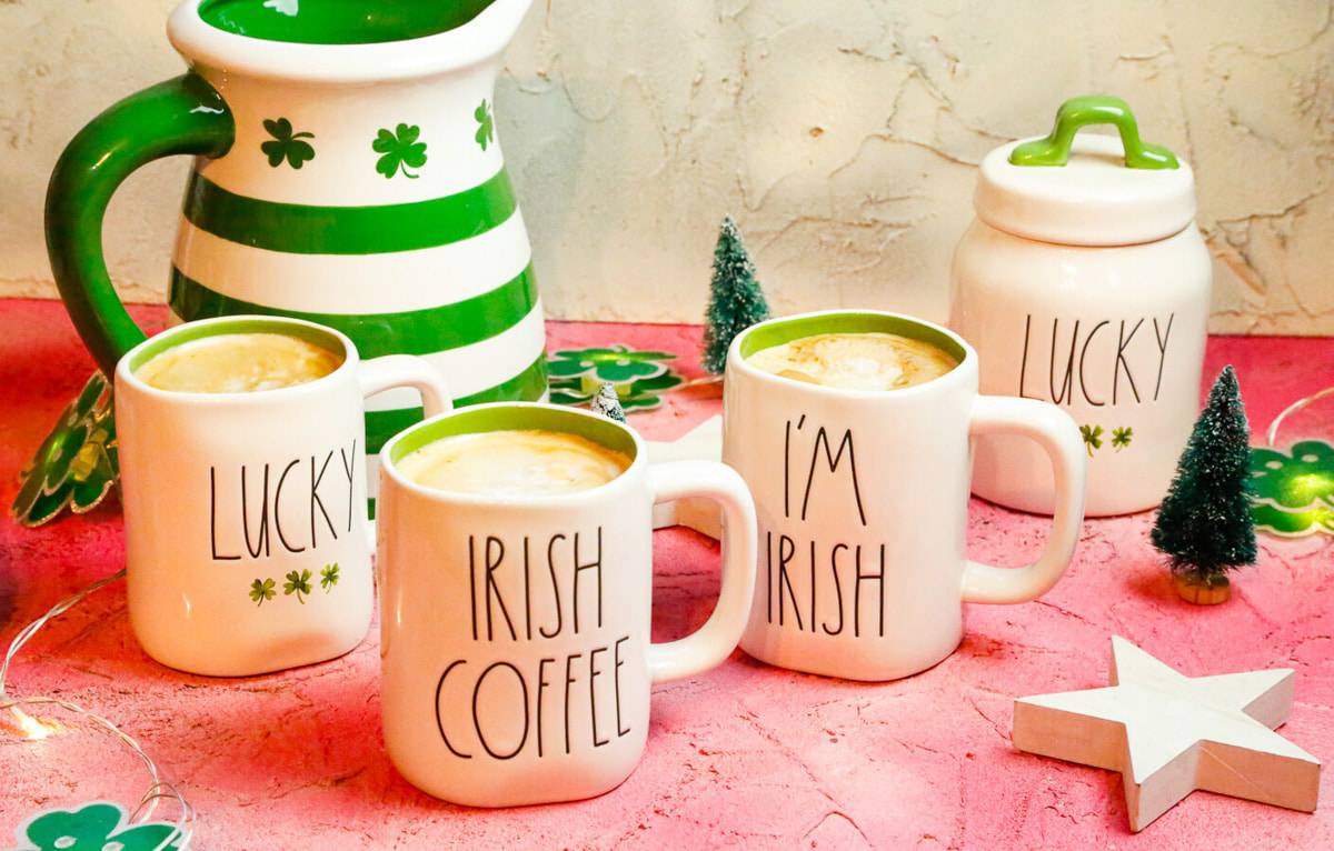 three coffee mugs filled with coffee and milk and Irish Cream with a green and white Saint Patricks Day jug behind the coffee mugs alongside a Lucky canister on a pink backdrop 
