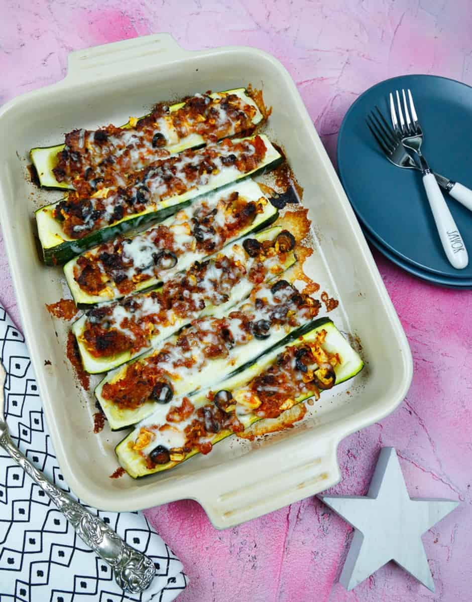 a dish of stuffed zucchini boats topped with melted cheese in a casserole dish on a pick back drop with a white star, a black and white napkin, blue plates and white forks 
