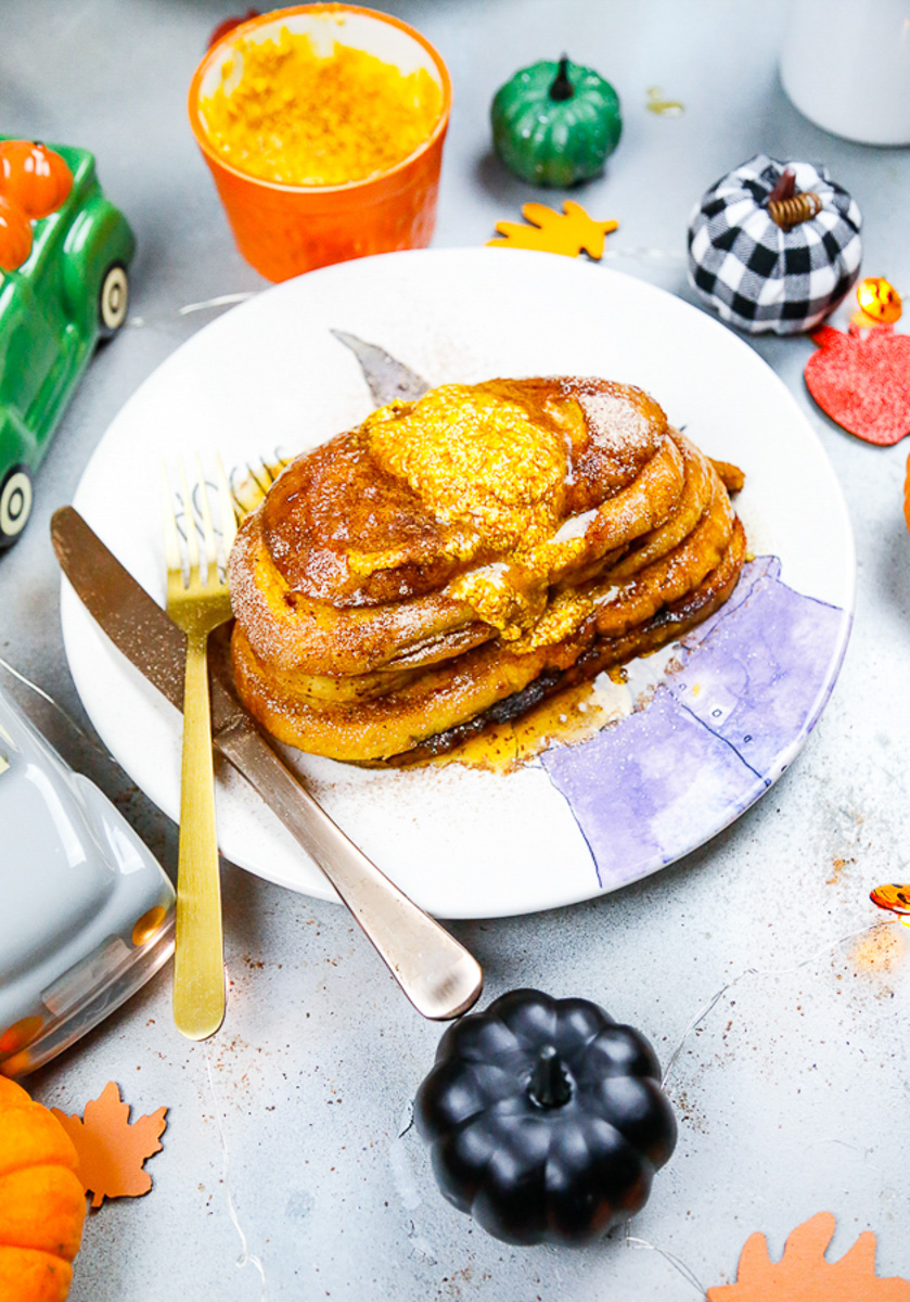 a stack of pumpkin brioche french toast with homemade pumpkin butter melted on top with a hocus pocus plate with a fork and knife and a bowl of pumpkin puree
