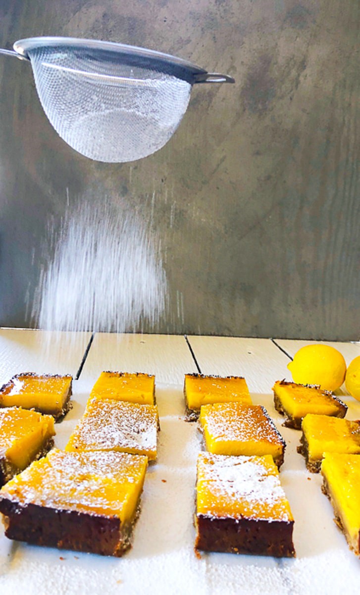 Power sugar being dusted on lemon bars with a grey background on a white wooden back drop 