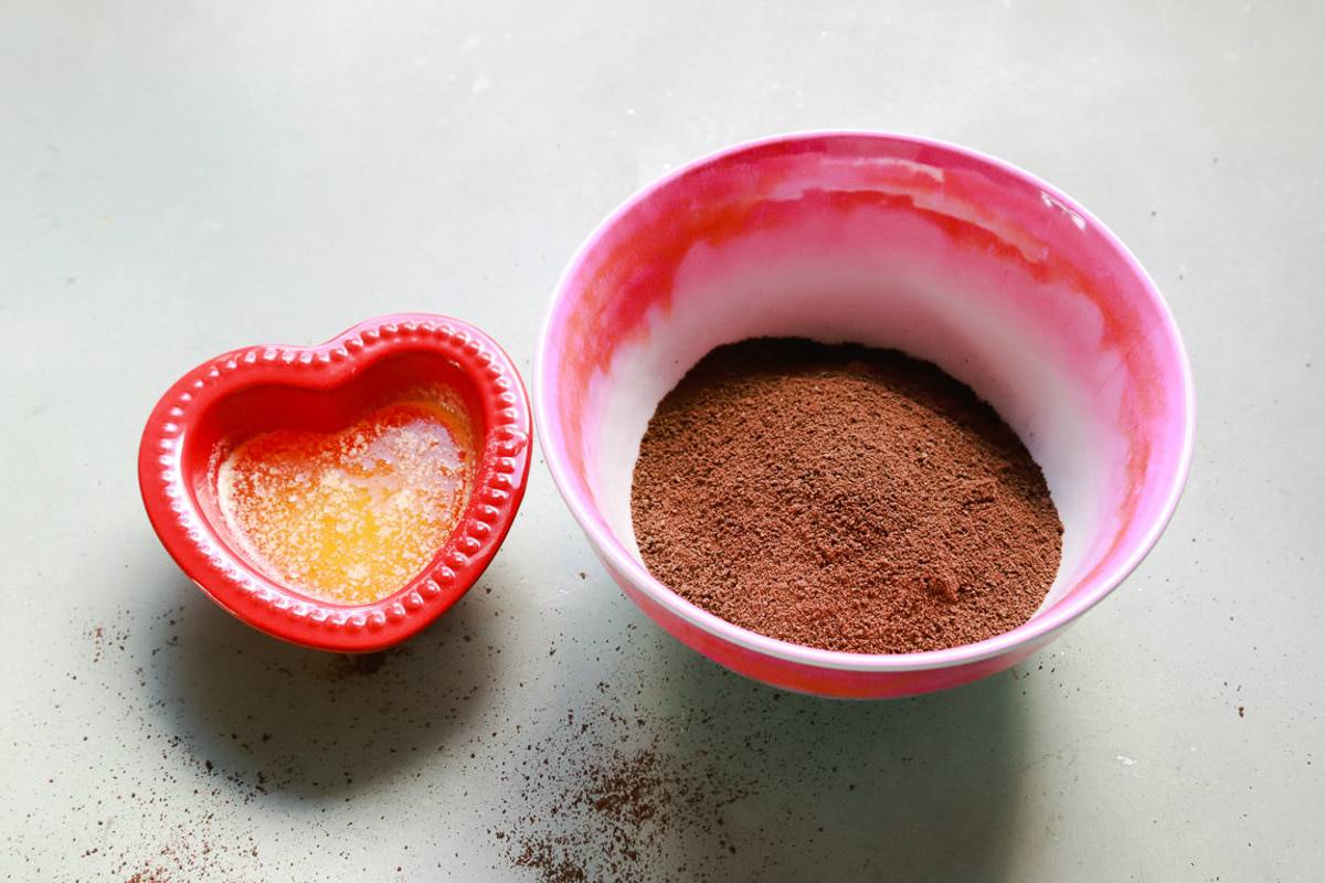 a red heart shaped bowl of melted butter and a pink and white bowl from Sur La Table with chocolate graham cracker crumbs and cocoa powder 