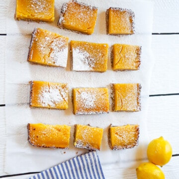 lemon bars sitting on a white wood backdrop with two lemons and a blue and white striped towel