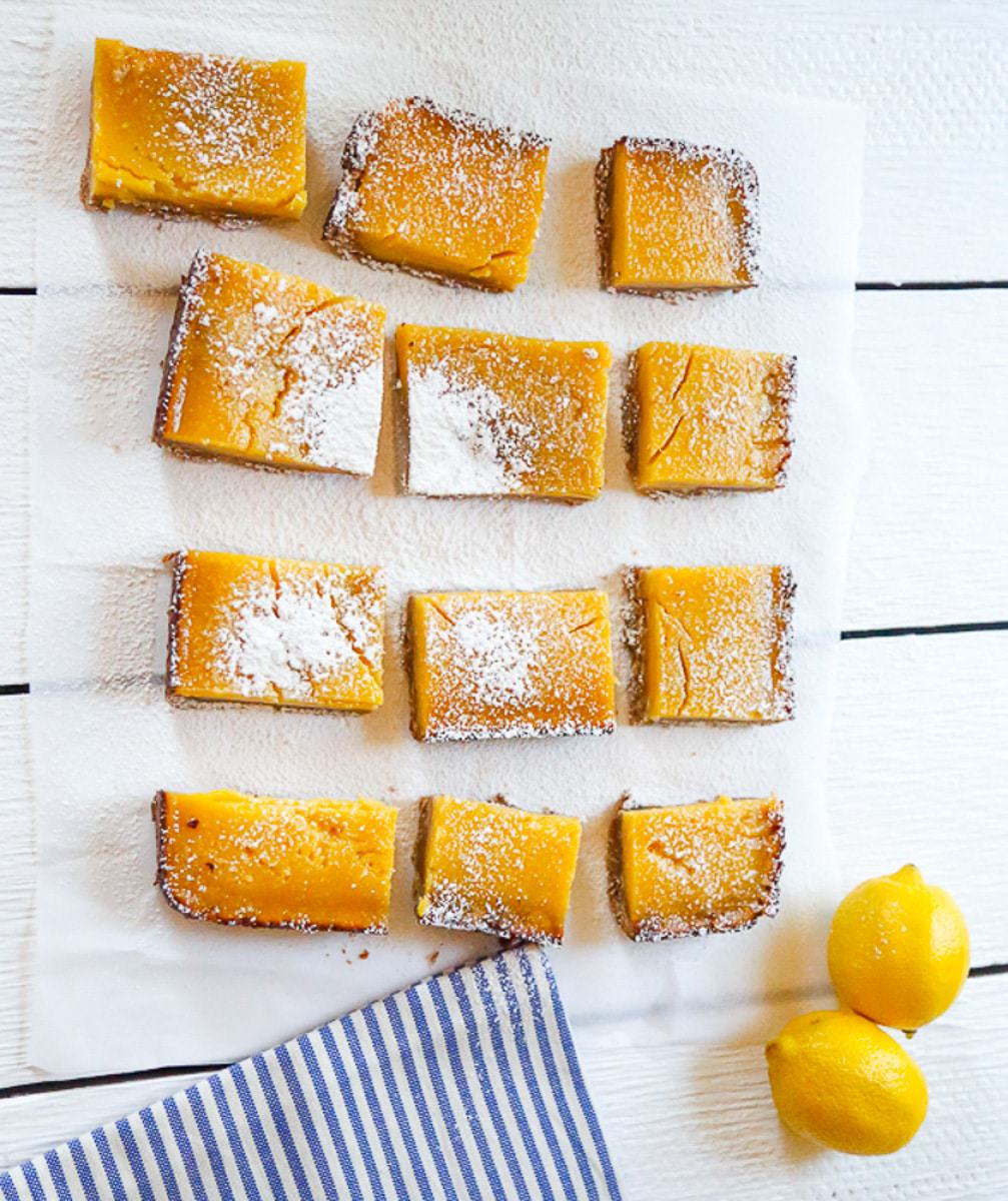 lemon bars sitting on a white wood backdrop with two lemons and a blue and white striped towel 