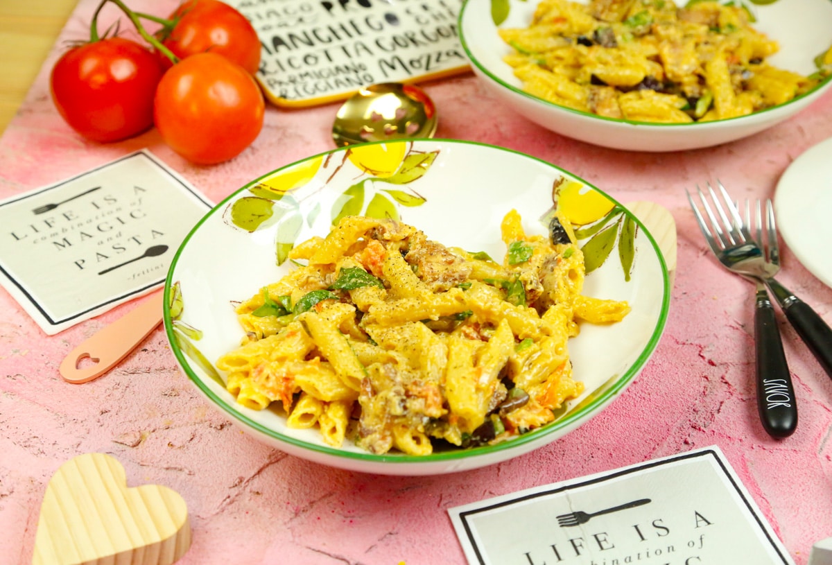 a bowl of cheesy penne pasta with tomatoes forks and napkins on a pink backdrop 
