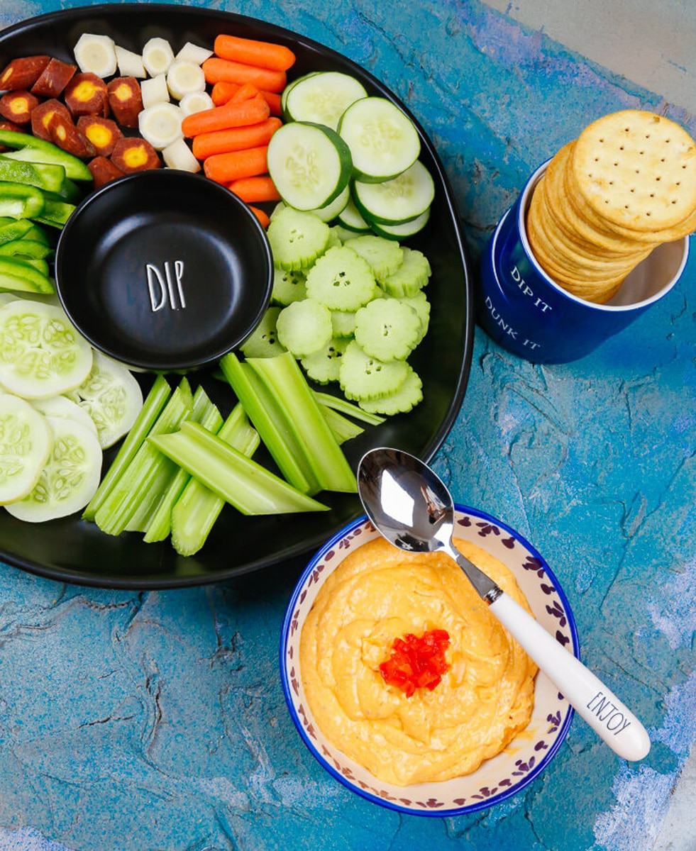 A dip tray filled with fresh veggies carrots, celery, cucumbers, lemon cucumbers, crackers, a spoon that says yum with a bowl of pimento cheese dip with pimentos on top 