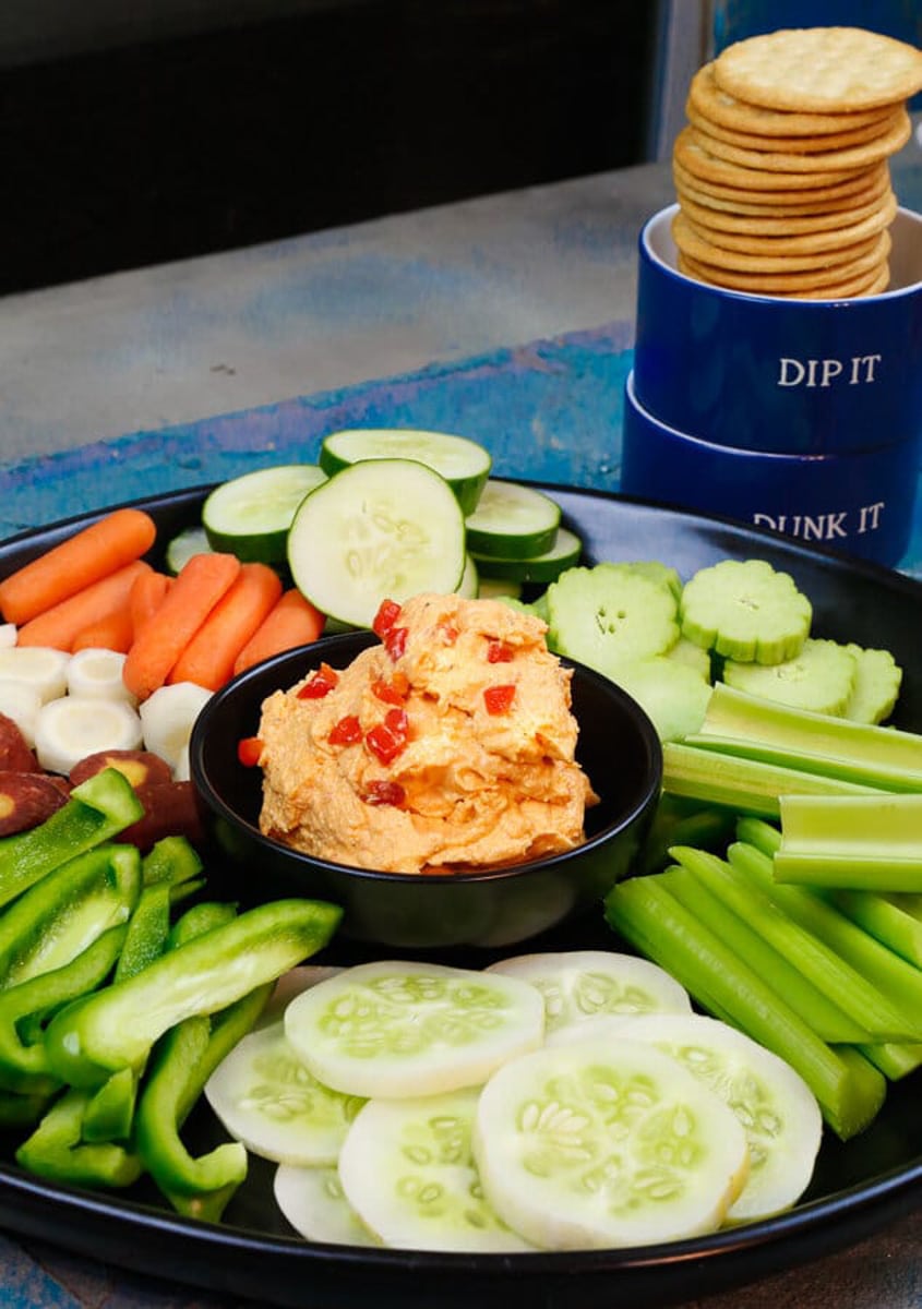A stack of crackers on a blue backdrop with vegetables and pimento cheese dip 