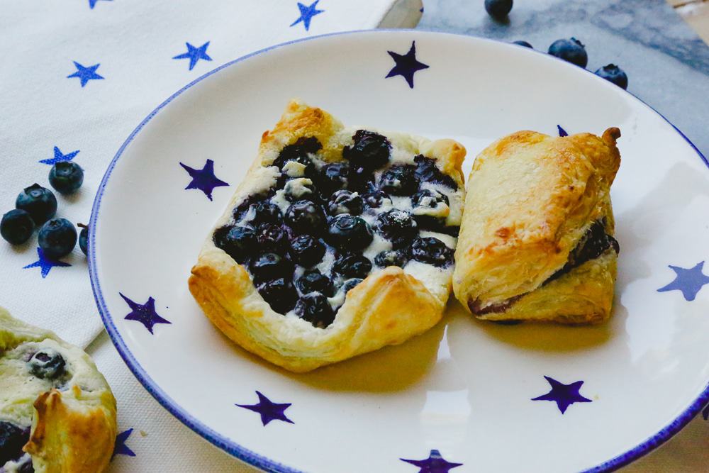 puff pastry blueberry turnovers sitting on a blue and white star plate with raw blueberries in the background on a marble backdrop 