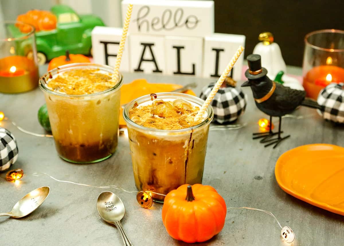 two glasses filled with pumpkin iced coffee with a faux pumpkin and a hello fall sign with a crow and a silver spoon 