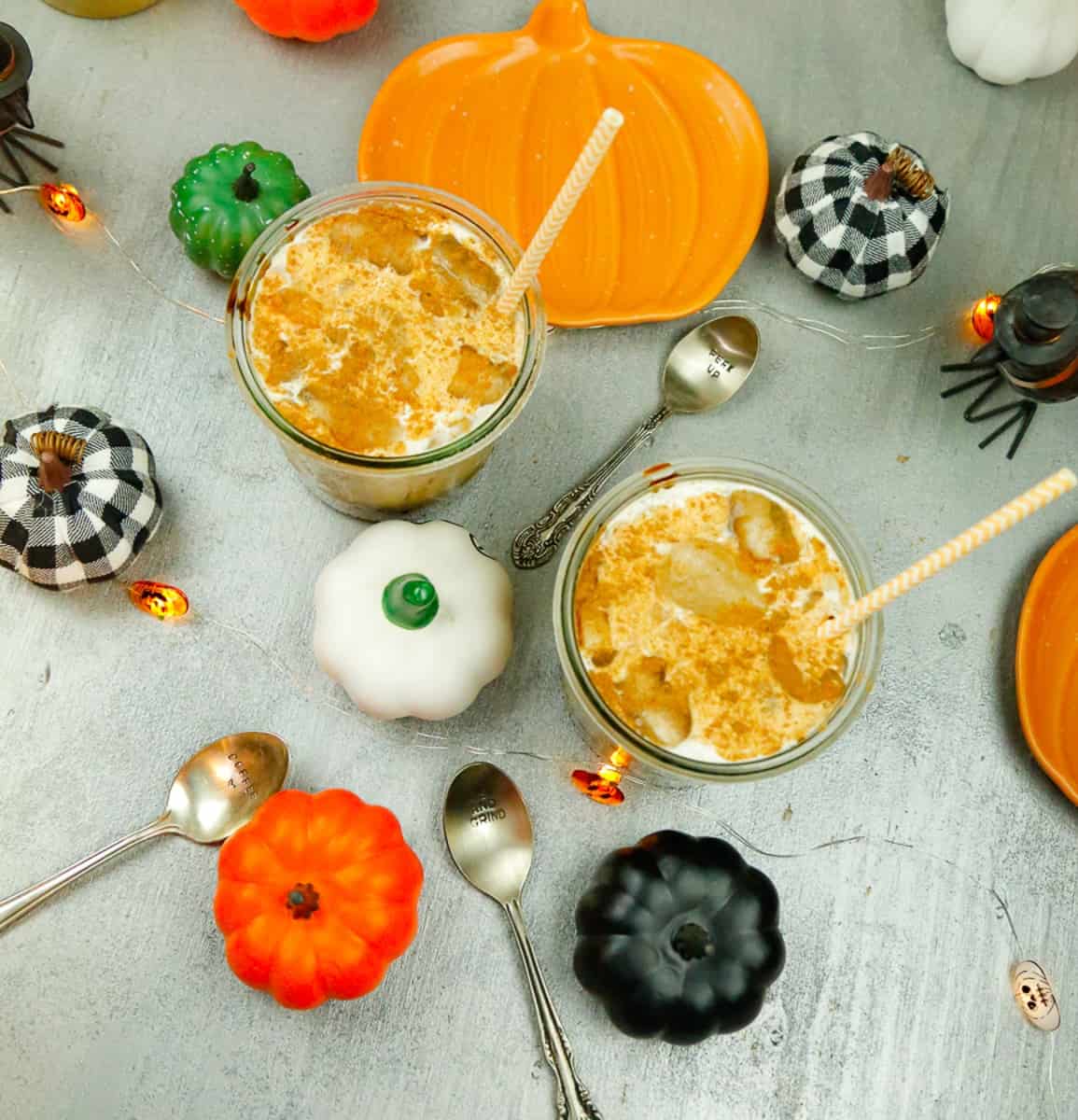 a flatlay of glasses filled with pumpkin iced coffee, silver spoons, and faux pumpkins on a silver wooden backdrop 