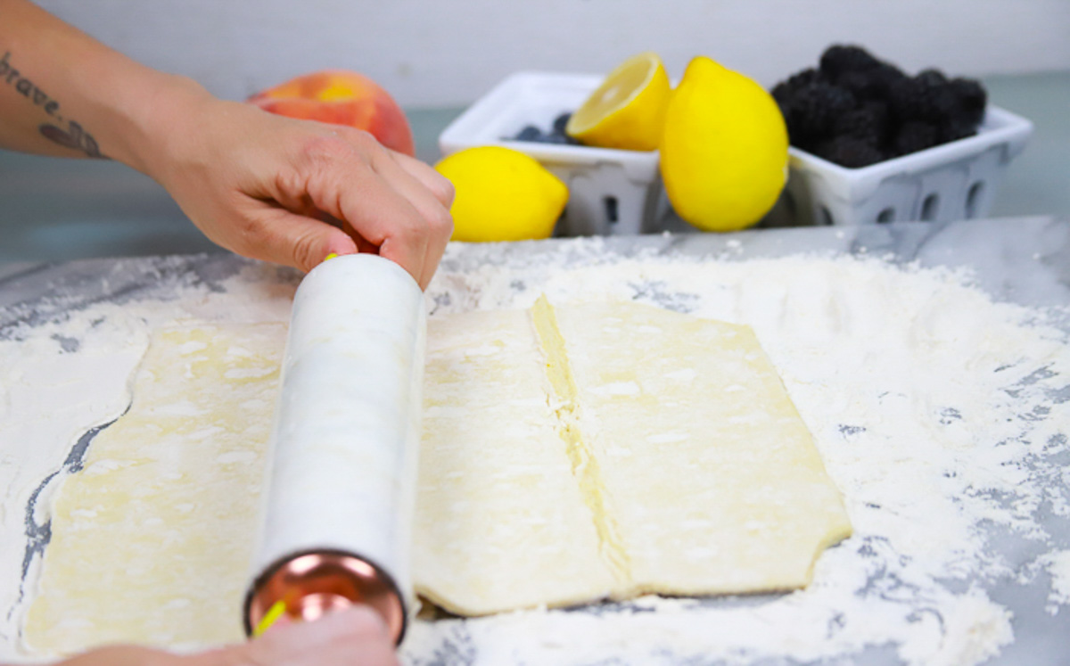 rolling pin rolling out thawed puff pastry dough on a floured surface with lemons and fresh berries 