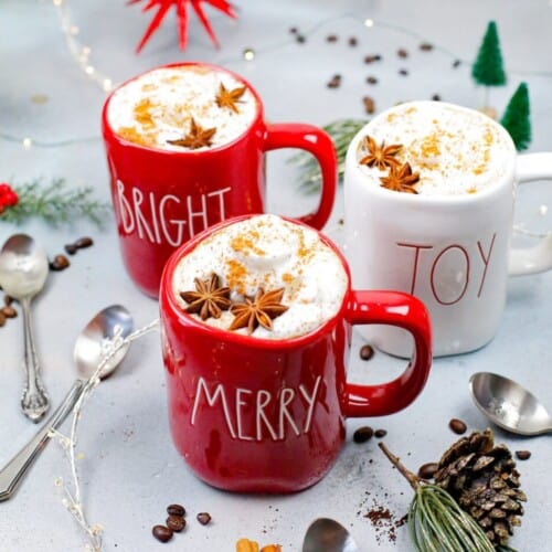 red and white mugs with the writing merry, joy, and bright on them full of coffee and steamed milk