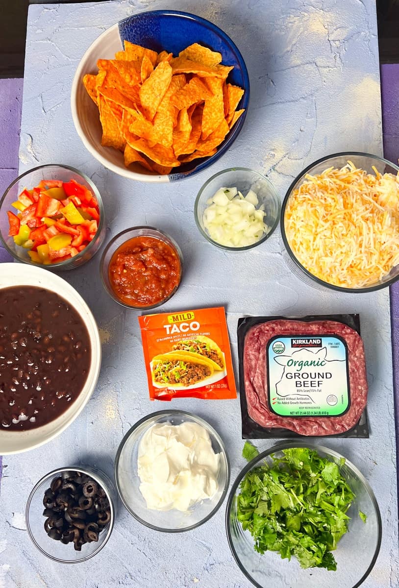 ingredients for Dorito casserole with ground beef, shredded cheese, taco seasoning, chopped onion, tomatoes, bell peppers, cilantro, black beans, sour cream, and nacho cheese Doritos 
