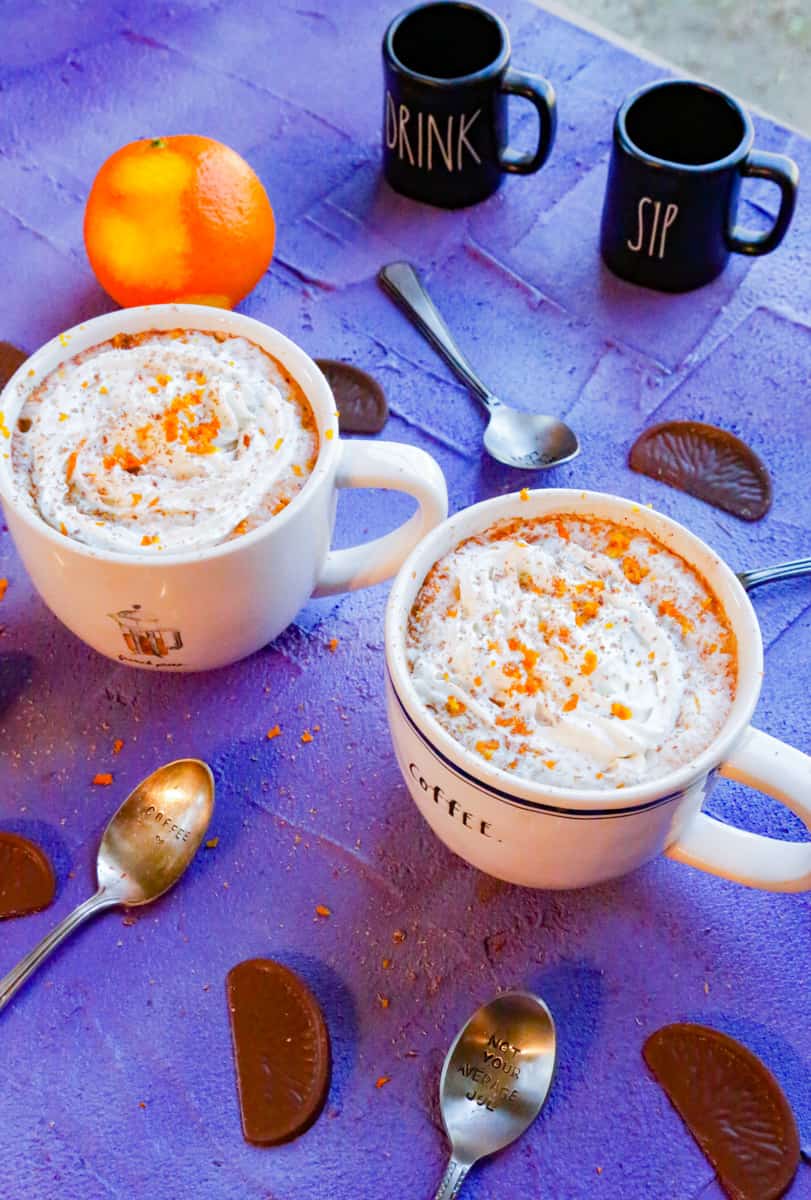 two cups of orange hot chocolate with whipped cream and orange zest along with chocolate orange slices and silver spoons on a purple backdrop