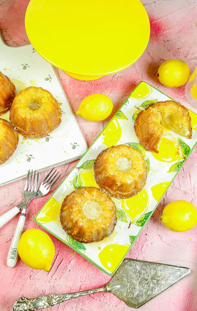 mini lemon bundt cakes on a lemon platter next to a yellow cake stand with fresh lemons and silverware