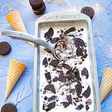 A tray of homemade oreo ice cream with a metal ice cream scoop and ice cream cones and oreos on a blue backdrop
