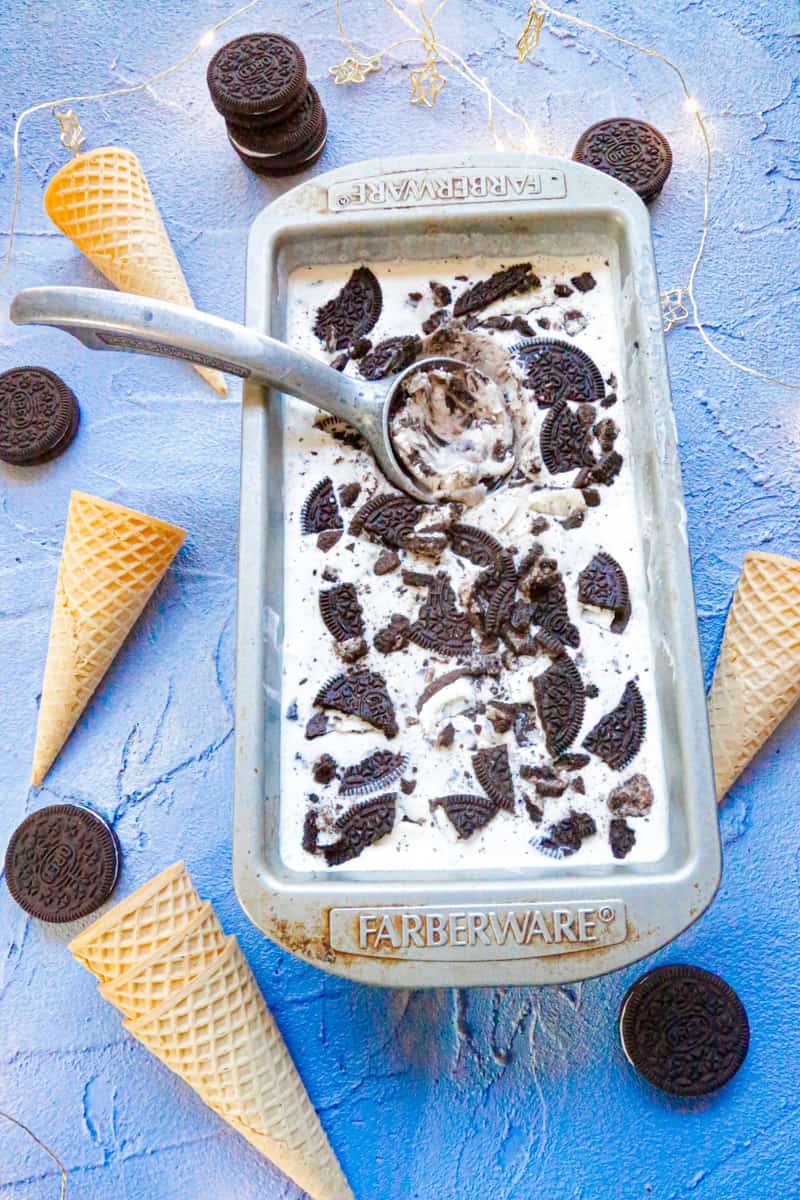 A tray of homemade oreo ice cream with a metal ice cream scoop and ice cream cones and oreos on a blue backdrop