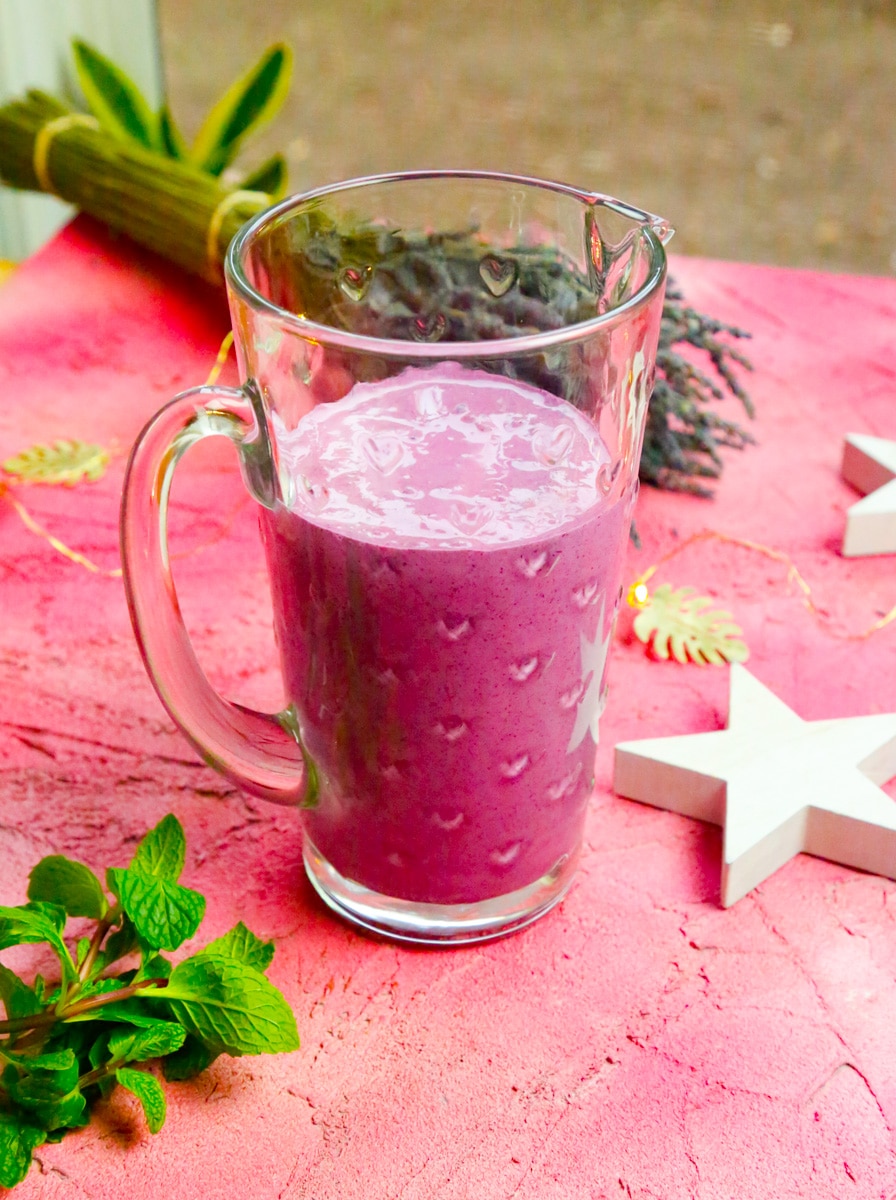 a pitcher of healthy blueberry milkshake with white stars and a bunch of mint with lavender in the background 
