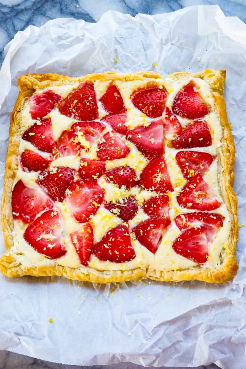 a baked strawberry puff pastry with lemon zest, cream cheese, on parchment paper on a marble back drop 