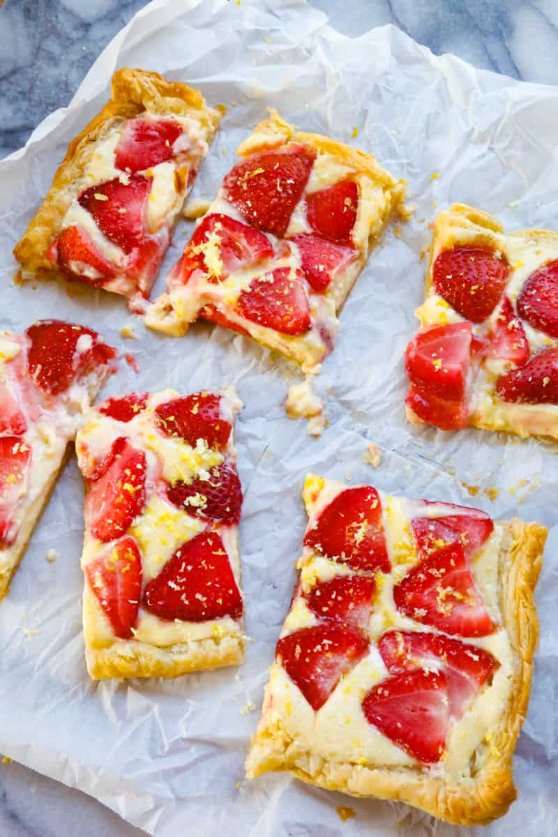 sliced strawberry turnovers with puff pastry and cream filling with lemon zest and parchment paper on a marble back drop. 