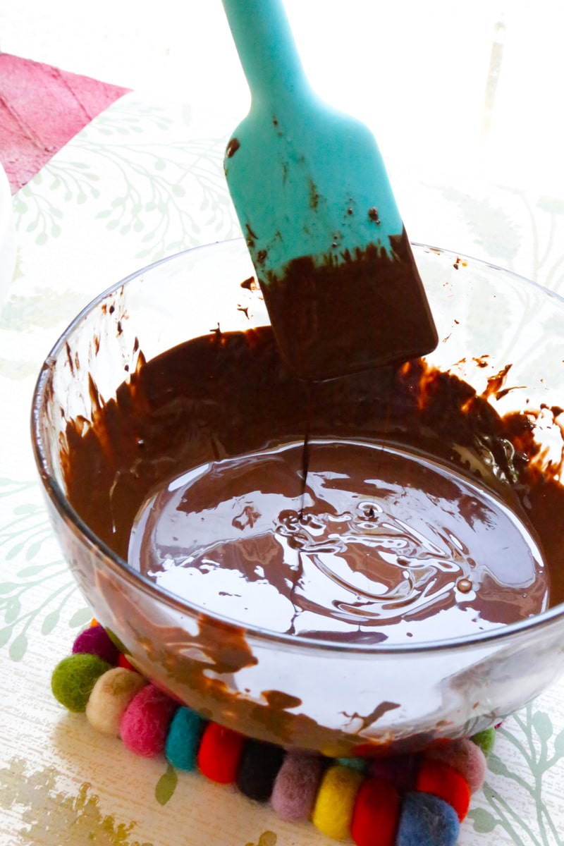 melted chocolate in a glass bowl with a blue spatula on a kitchen table sitting on a multicolored heat mit 