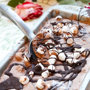 a silver cake tray of chocolate marshmallow ice cream drizzled with chocolate sauce and marshmallows with a silver ice cream scoop on a backdrop with flowers