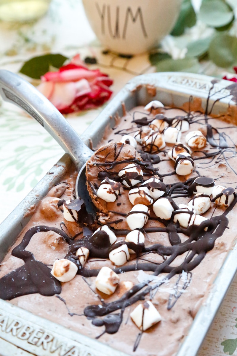 a silver cake tray of chocolate marshmallow ice cream drizzled with chocolate sauce and marshmallows with a silver ice cream scoop on a backdrop with flowers 