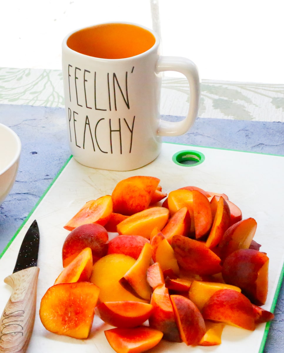 sliced peaches on a white cutting board with a knife that reads slice and a Rae Dunn mugs that says Feelin Peachy 