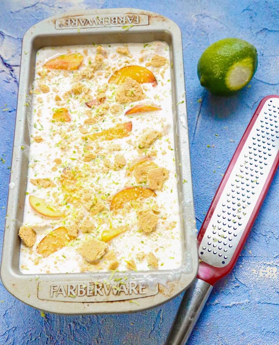 lime zest on peaches and cream ice cream in a metal loaf pan with a lemon zester and a lime on a blue back drop 