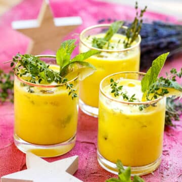 two glasses of pineapple and gin juice with fresh sprigs of mint and rosemary two stars and fresh lavender sprigs on a pink backdrop