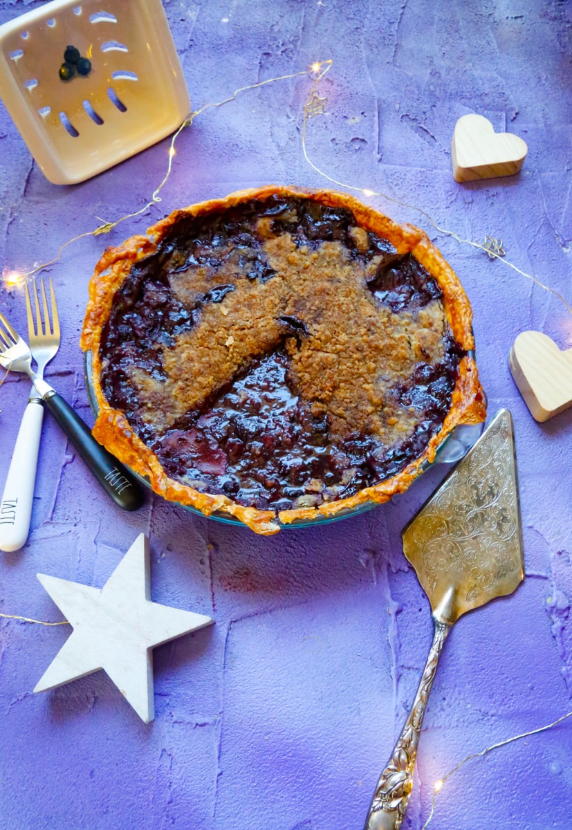 delicious blackberry crumble pie with a sterling silver pie server, decorative stars, two Rae Dunn forks, on a purple back drop with a blueberry basket 