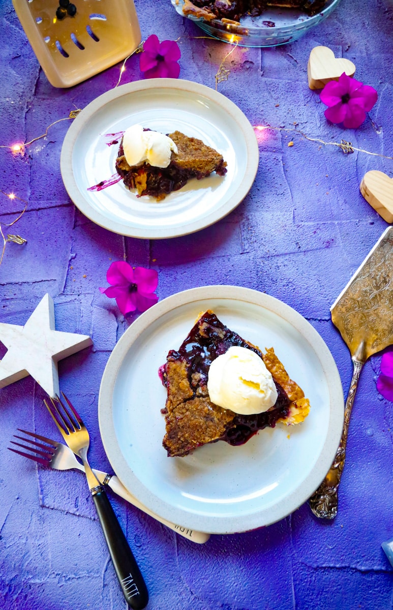 two slices of berry pie topped with vanilla ice cream with purple petunias and white stars on a purple back drop with forks and a silver pie server 
