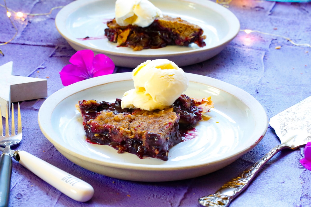 two slices of blackberry pie topped with a scoop of vanilla ice cream with forks, stars, and a metal pie server on a purple back drop 