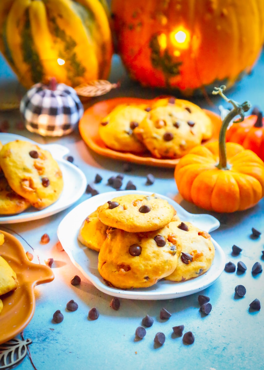 a pumpkin plate of chewy pumpkin cookies with chocolate chips around it and pumpkins in the background 