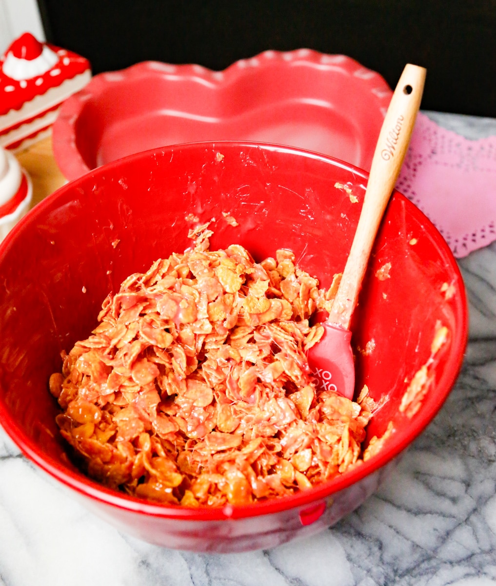 crispy cornflake cookie batter in a large red mixing bowl with a cookie batter spoon and a heart shaped pie pan. 
