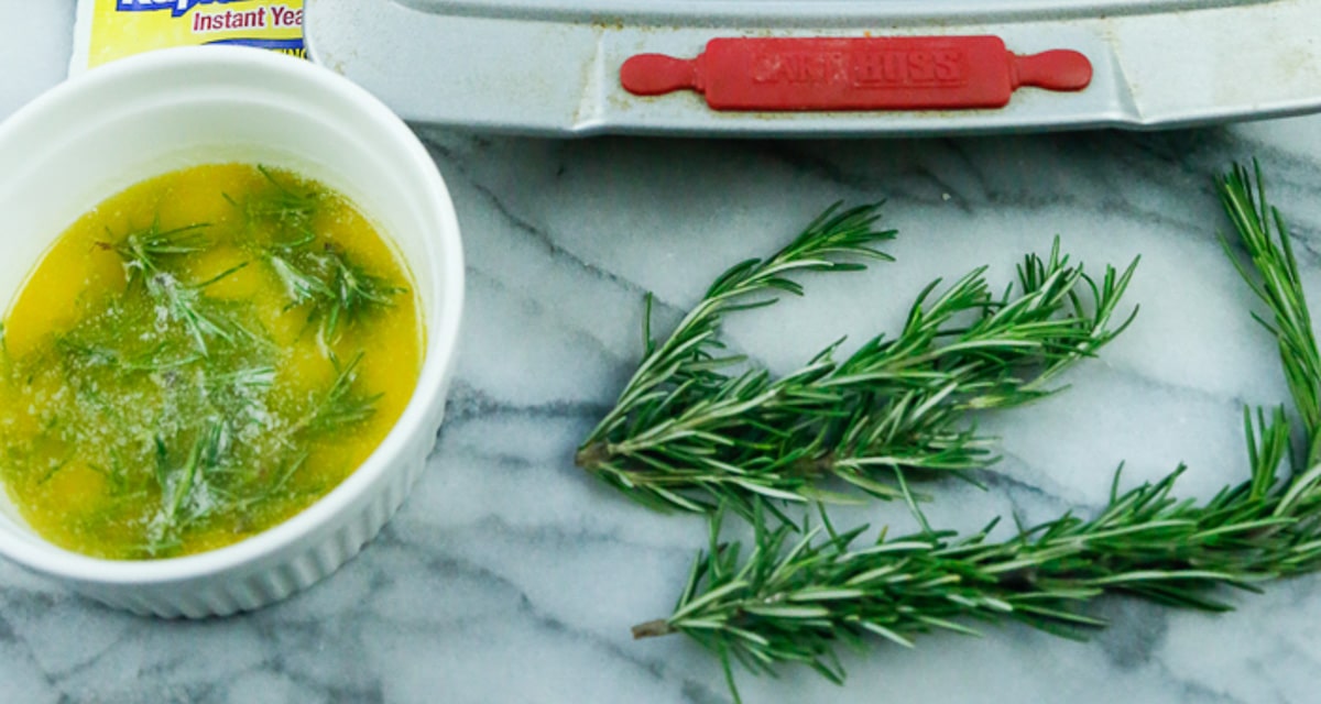 A bowl of fresh rosemary butter with fresh rosemary sprigs on a marble backdrop for buttery buns 