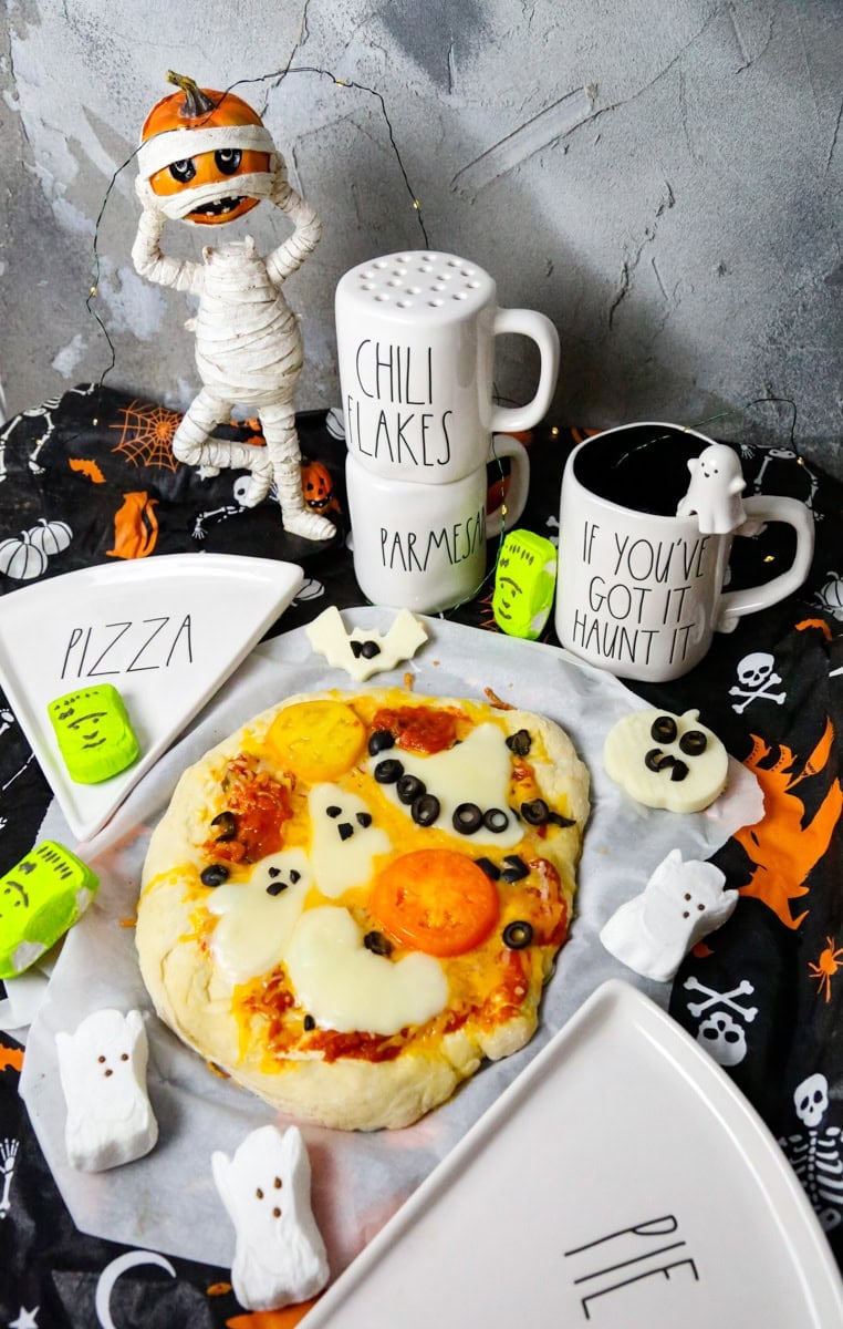 Spooky pizza with spooky mozzarella ghosts and a witch hat and a bat on a Halloween tablecloth with a Jack-o-latern mummy holding his head with pizza and pie pizza plates 
