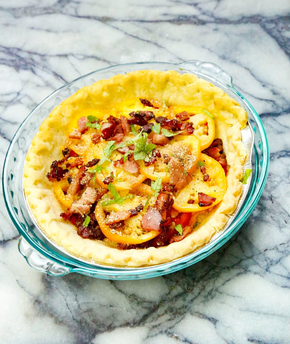Now it's time to assemble the rest of our tomato and cheese pie. In alternating layers, fill pie crust with tomatoes, bacon, green onions, chives, and fresh basil, and garlic powder, and fresh black pepper.