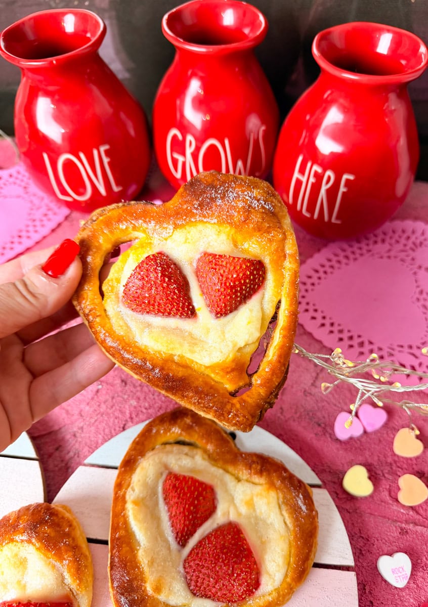 Hand holding a heart shaped strawberry cream cheese danish made with puff pastry, topped with fresh strawberries, with Valentine’s decor in the background