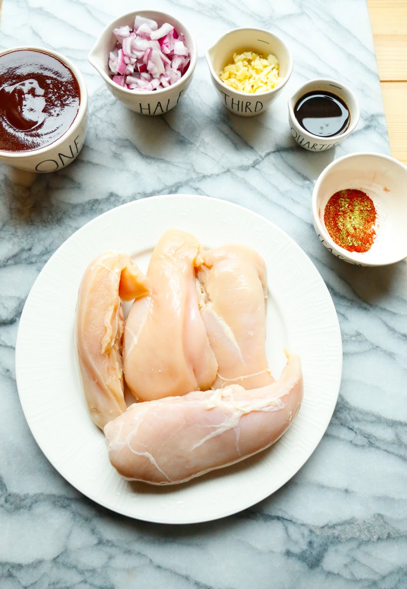 Ingredients for barbecue chicken sandwich slow cooker recipe including raw chicken breasts, diced onion, garlic, barbecue sauce, molasses, and spices on a marble surface.