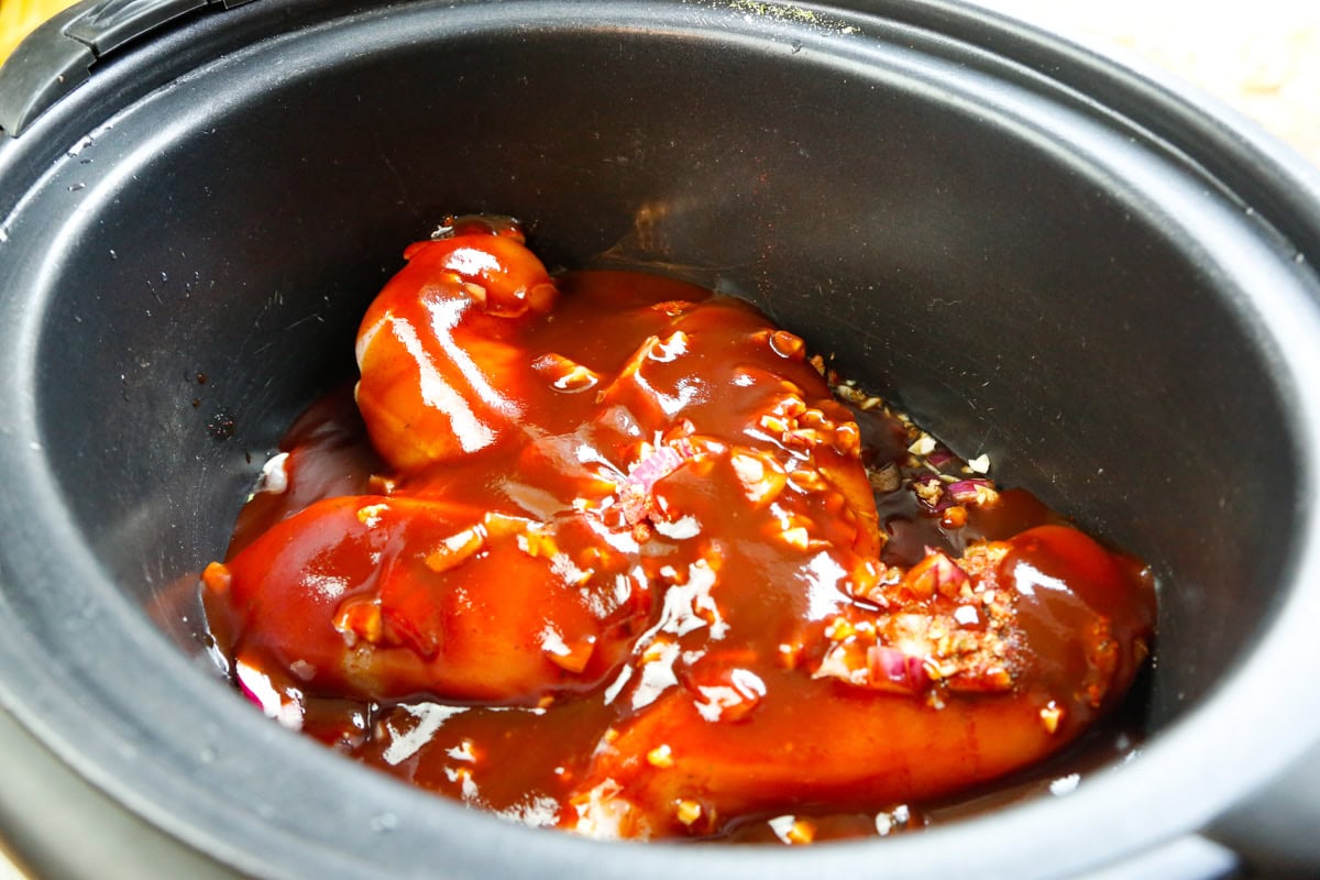 Barbecue sauce poured over chicken breasts, garlic, and red onion in a slow cooker for shredded BBQ chicken sandwiches