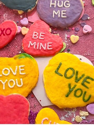 Colorful conversation hearts sugar cookies decorated with royal icing and phrases like “Love You,” “Be Mine,” and “Hug Me” on a pink Valentine’s Day background.