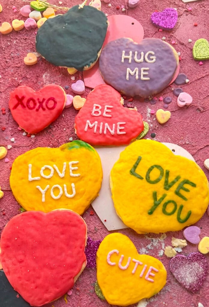 Frosted conversation heart sugar cookies with royal icing sayings including “XOXO,” “HUG ME,” and “BE MINE,” styled for a Valentine’s Day cookie recipe.