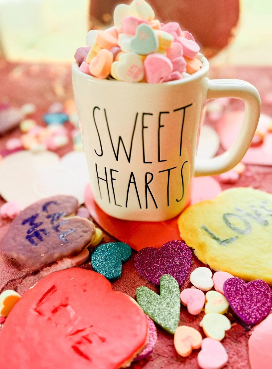 Rae Dunn “Sweet Hearts” mug surrounded by decorated conversation hearts sugar cookies and colorful Valentine’s Day candy on a pink background