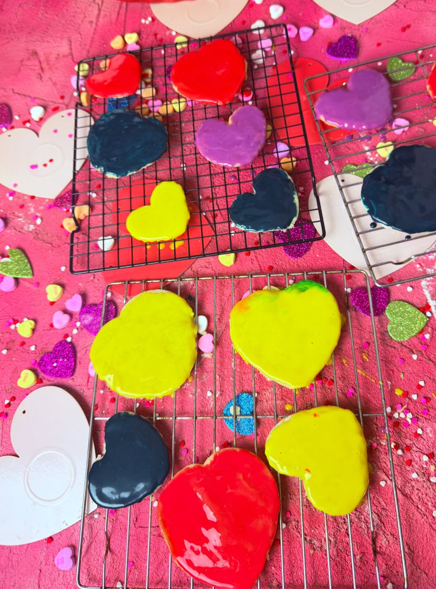 Colorful heart shaped sugar cookies decorated with royal icing in bright yellow, red, purple and black on cooling racks for Valentine’s Day conversation hearts cookies